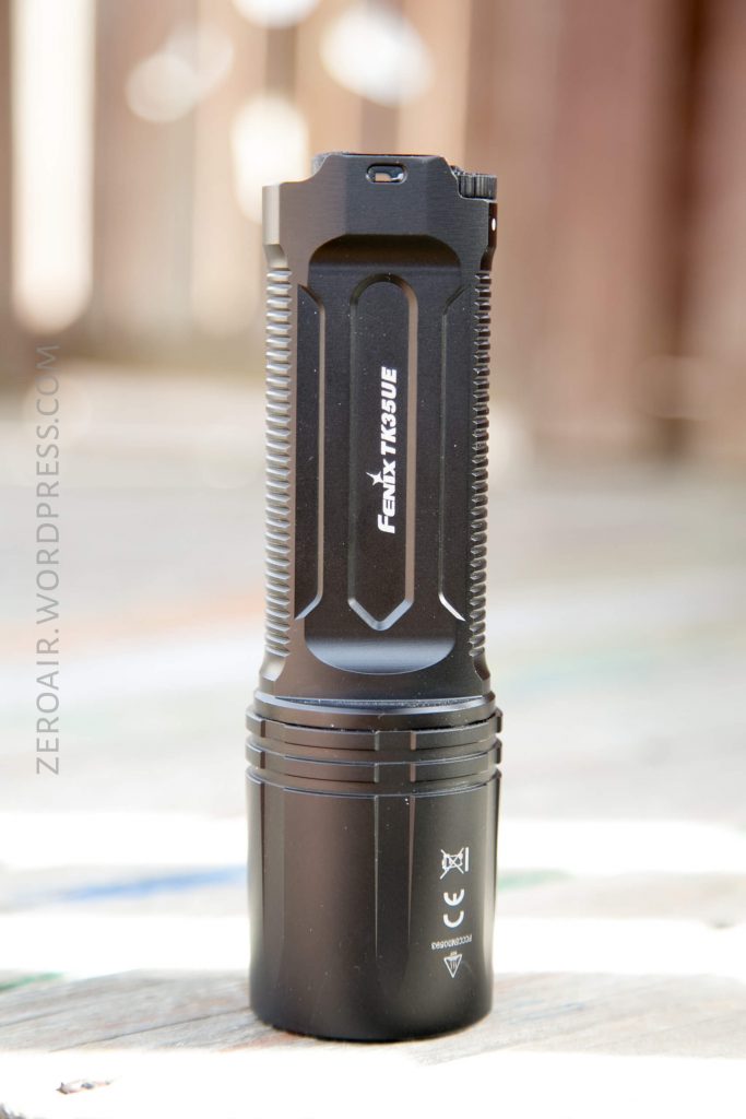 A black Fenix TK35UE flashlight stands upright on a flat surface, with textured grip and logos visible on the body. The background is blurred and out of focus.