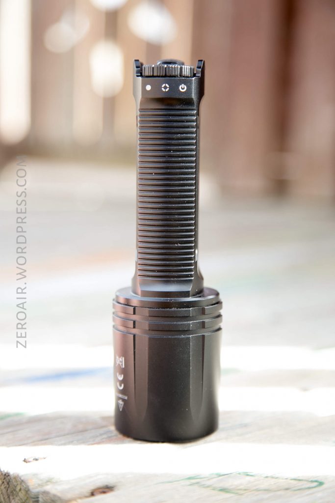 A black, ribbed handheld flashlight is standing upright on a wooden surface, with a blurred outdoor background. The side of the flashlight displays some small white symbols.