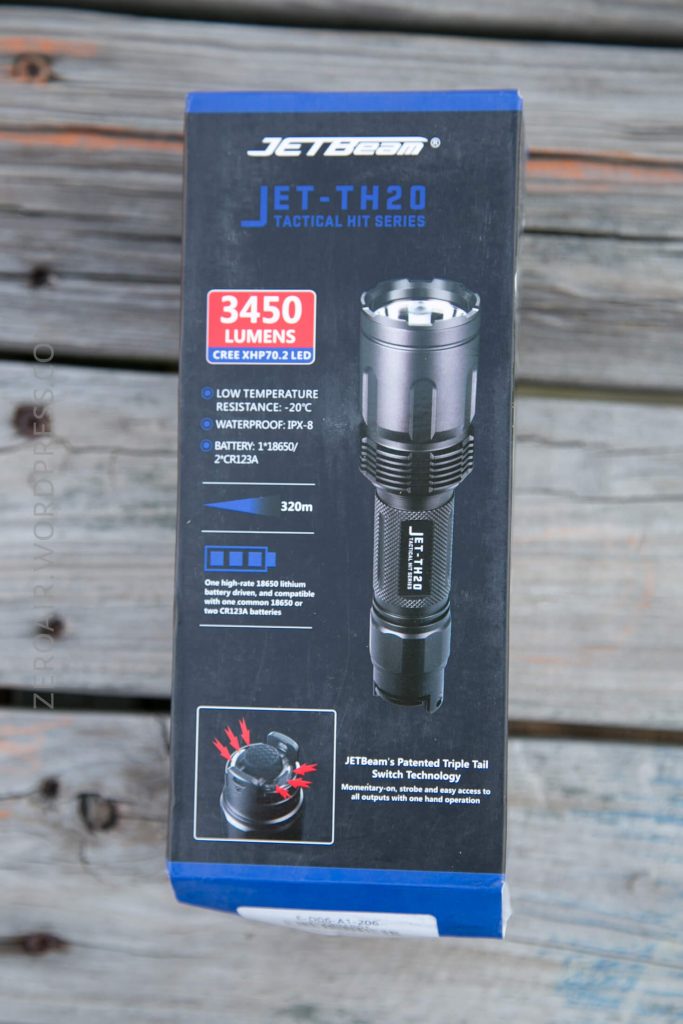 A product box for the JETBeam JET-TH20 tactical flashlight is on a wooden surface. The box highlights features such as 3450 lumens, IPX-8 waterproof rating, and a 320m range, with an image of the flashlight on the front.