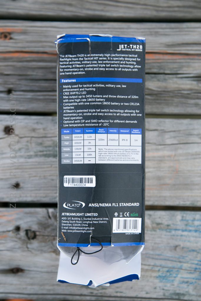 A product box for the JETBeam TH20 tactical flashlight displays features, specifications, certification marks, and company contact information against a wooden background. The box is slightly damaged at the bottom.