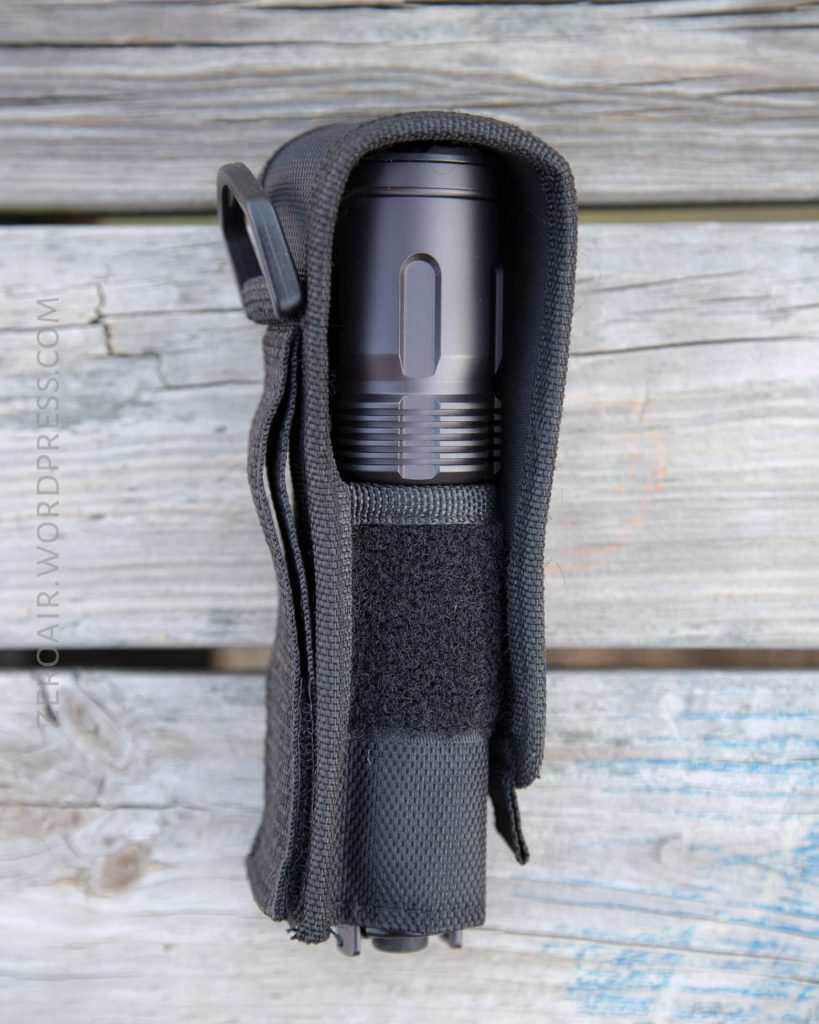 A black tactical flashlight is secured in a fitted black nylon holster with a Velcro flap, resting on a weathered wooden surface.