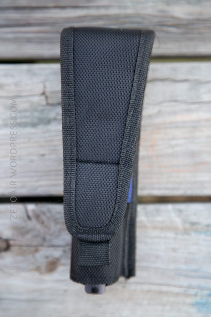 A black textured nylon holster with a velcro flap is standing upright on a weathered wooden surface. The side seam and a small portion of a blue object inside are visible.