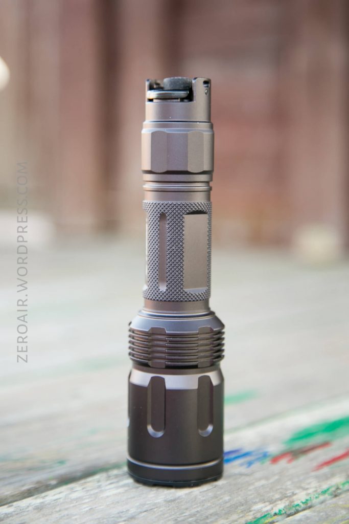 A grey, metallic flashlight stands upright on a wooden surface with a blurred brown background. The flashlight has textured grip sections and a button on the top.