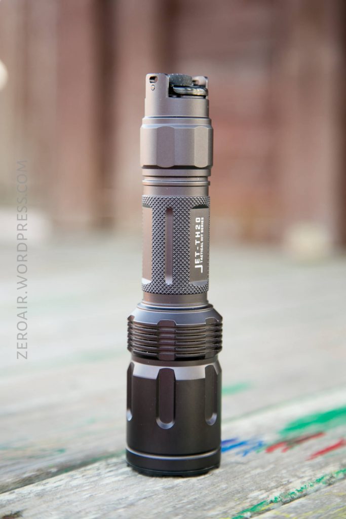 A metallic, compact flashlight stands upright on a weathered wooden surface with some colorful markings. The brand name “JETBeam” is visible on the side. The background is blurred.