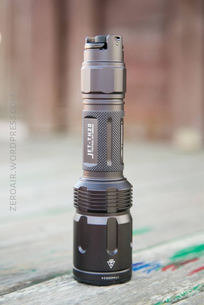 A black JETBeam tactical flashlight stands upright on a weathered wooden surface with faint, colorful markings. The background is blurred, and the flashlight details and branding are clearly visible.
