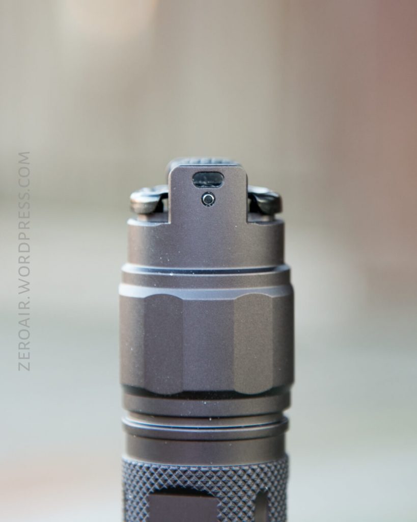 A close-up view of the top part of a metallic flashlight with a black finish, showing a button and textured grip. The background is blurred. The text ZEROAIR.WORDPRESS.COM appears vertically on the left side.