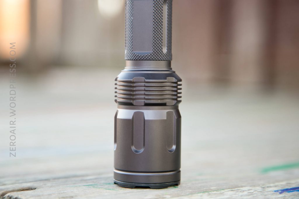A close-up view of a metallic flashlight standing upright on a wooden surface, showing the textured grip and details of the flashlight body. The background is blurred.
