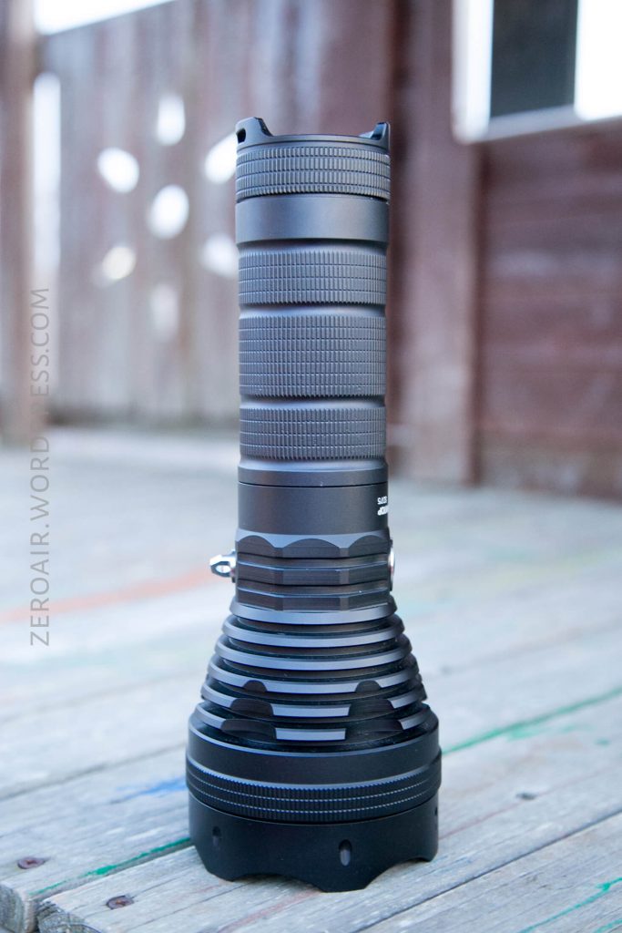 A large, black tactical flashlight is standing upright on a wooden surface outdoors. The background is blurred, showing part of a wooden structure with windows. The flashlight has a ribbed grip and wide head.