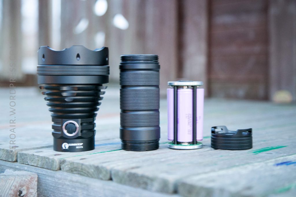 A disassembled flashlight is displayed on a wooden surface, showing four main components: the lamp head, battery tube, battery holder with two cylindrical batteries, and the tail cap. The background is blurred.