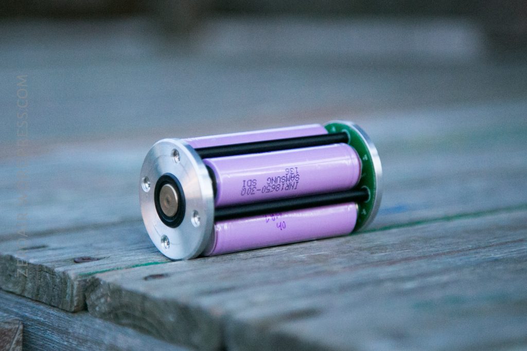 A cylindrical battery holder containing three pink rechargeable lithium-ion batteries is placed on a wooden surface outdoors.