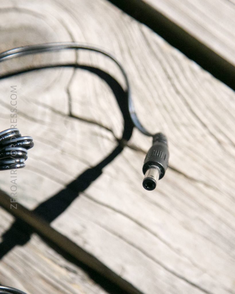 A close-up of a black power cable with a barrel connector resting on a wooden surface. The cable casts a shadow, and the wood grain is visible.