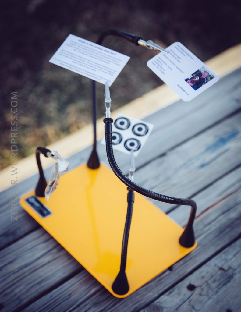 A yellow stand with three flexible arms holding cards and small objects, placed on a wooden surface. The arms have metal clips at the ends securing the items. The background is out of focus.