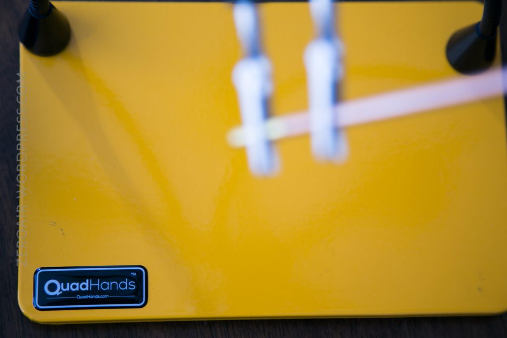 A close-up of a yellow QuadHands workbench with two blurred vertical clamps in the center and a visible QuadHands logo in the bottom left corner.