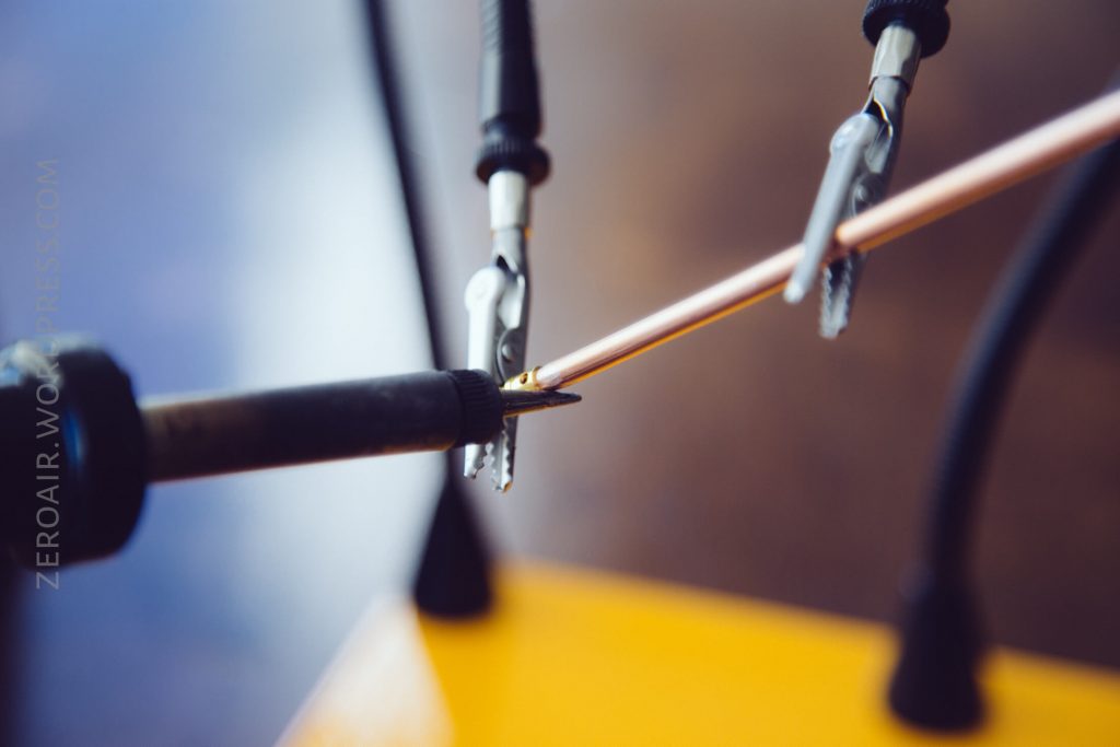 A close-up of a soldering iron tip touching a metal wire held by two small alligator clips, with a blurred yellow base and background.