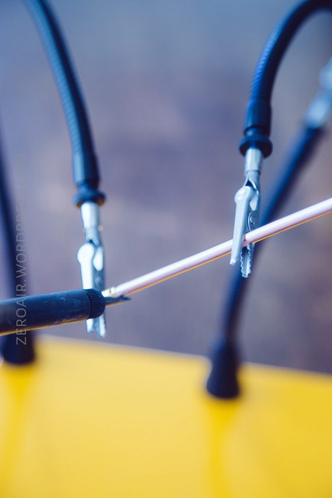 Close-up of three alligator clips attached to a thin white rod or wire, with blurred background and part of a yellow surface visible at the bottom of the image.