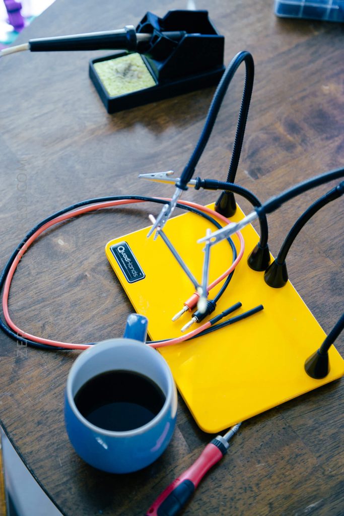 A yellow soldering mat with flexible helping hands holds wires on a wooden table. A soldering iron, tip cleaner, red-handled tool, and a blue mug of coffee are also visible.