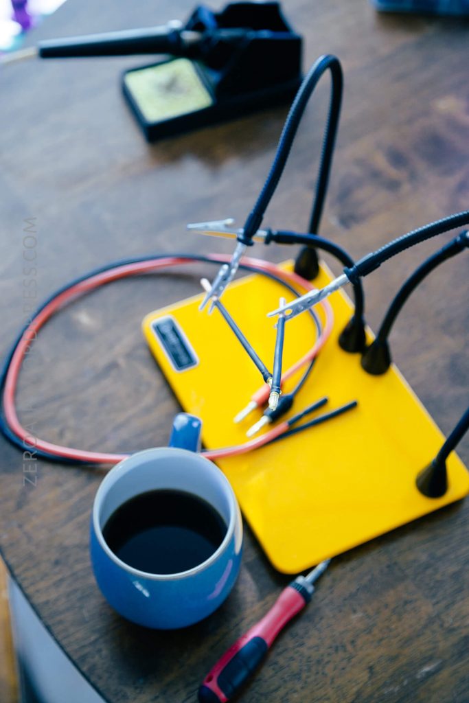 A yellow soldering mat with flexible helping hands holds wires on a wooden table. A cup of black coffee and a red screwdriver are in the foreground; a soldering iron stand is in the background.