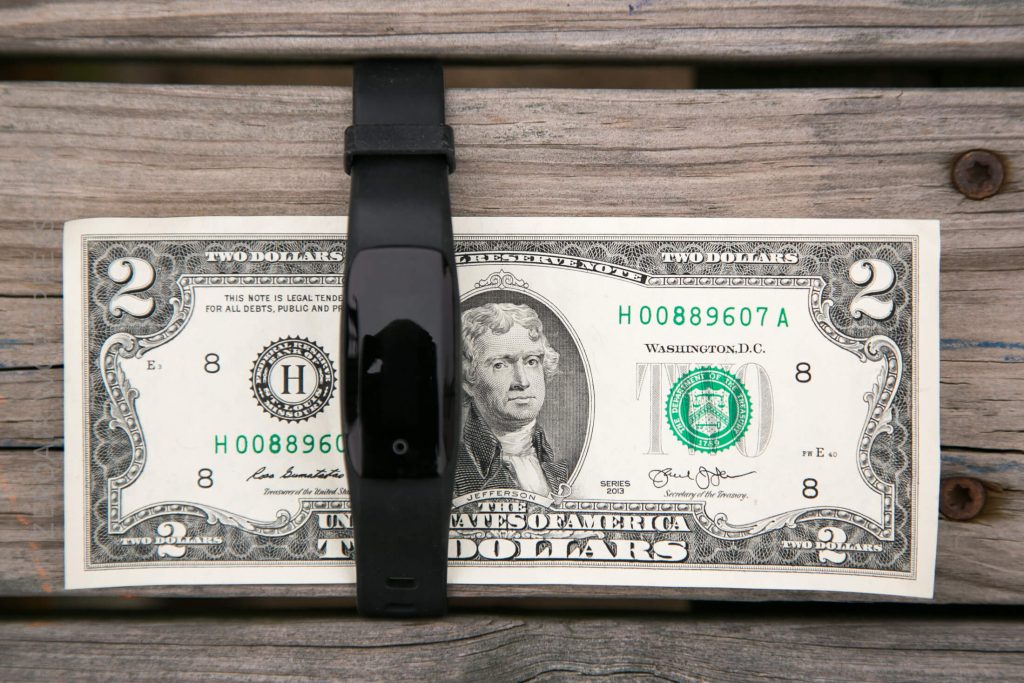 A two-dollar bill is held down on a wooden surface by a black fitness tracker placed horizontally across its center.