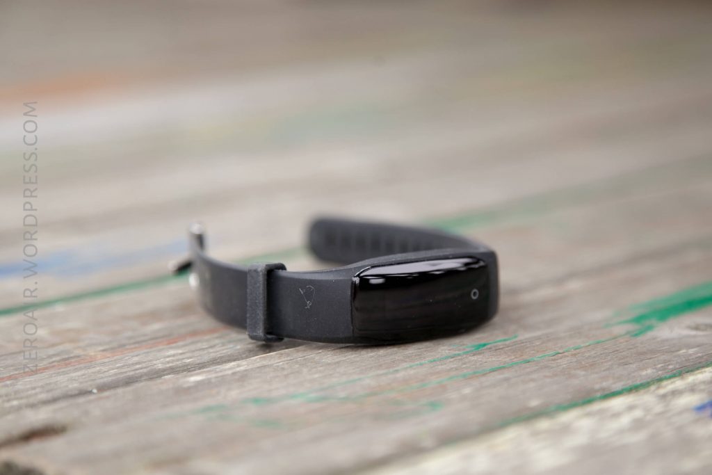 A black fitness tracker with a glossy screen and silicone strap rests on a weathered wooden surface. The background is blurred.