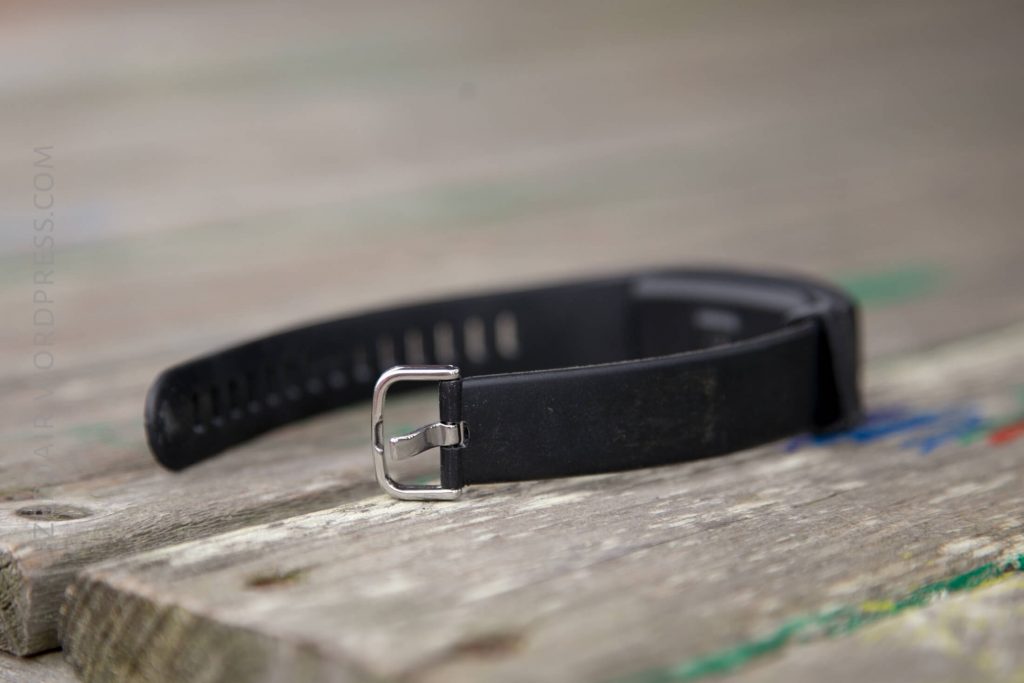 A black rubber watch strap with a silver buckle is placed on a weathered wooden surface, photographed at a slight angle.