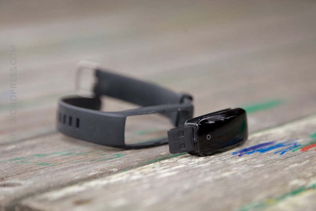 A black fitness tracker wristband lies on a wooden surface, with the tracker module detached and placed beside the empty band. The USB connector is visible on the module.