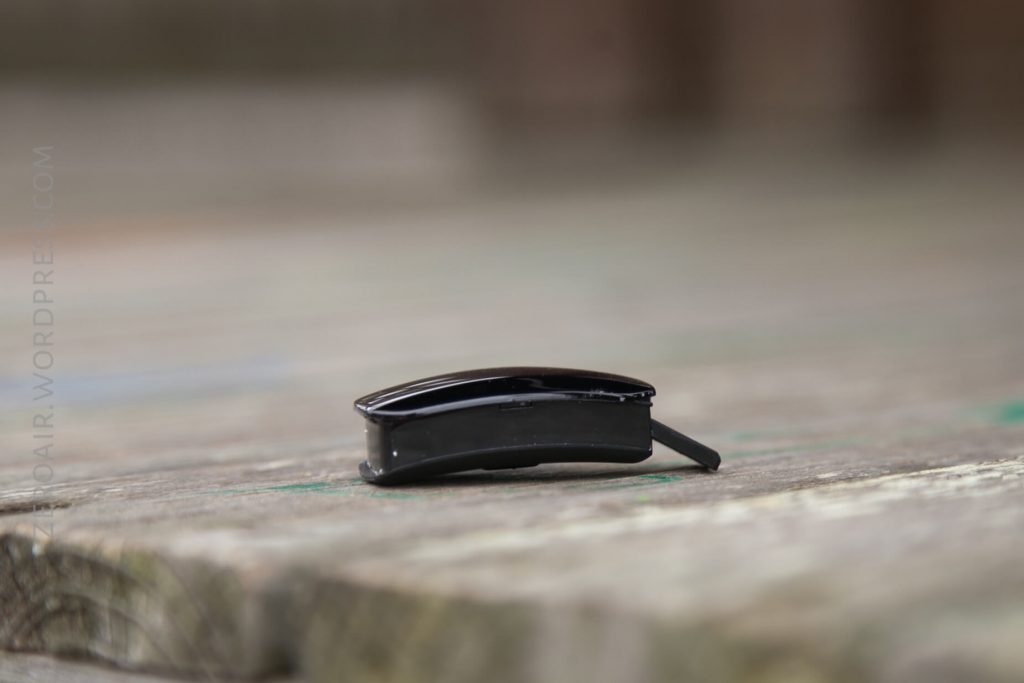 A small, black Bluetooth earpiece rests on a wooden surface. The earpiece is in focus while the background is blurred.