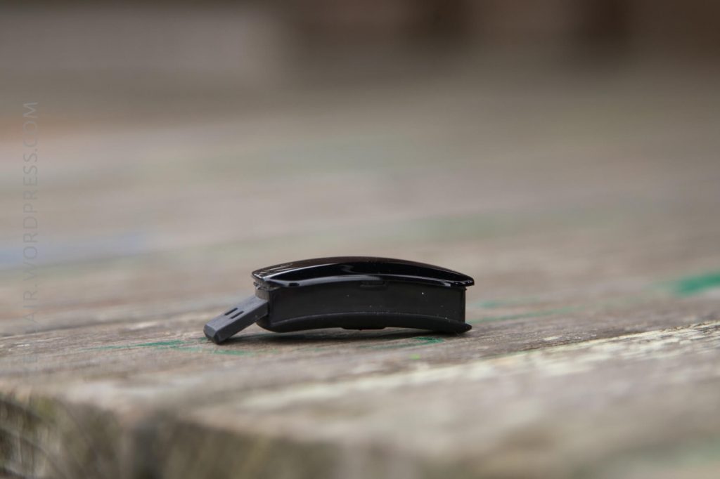 A small, black, curved plastic object with a clip, possibly a hairpin or a barrette, lying on a weathered wooden surface with green paint marks. The background is out of focus.