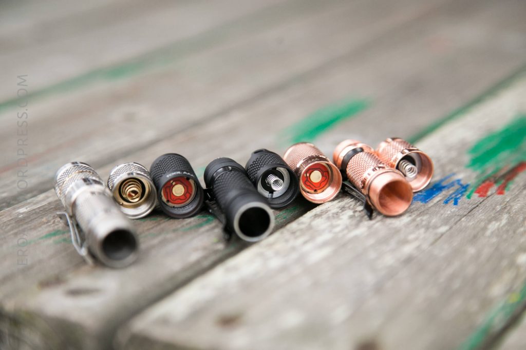 Five small cylindrical metal flashlights in various finishes, including silver, black, and copper, are lined up side by side on a weathered wooden surface with some green and red paint marks.