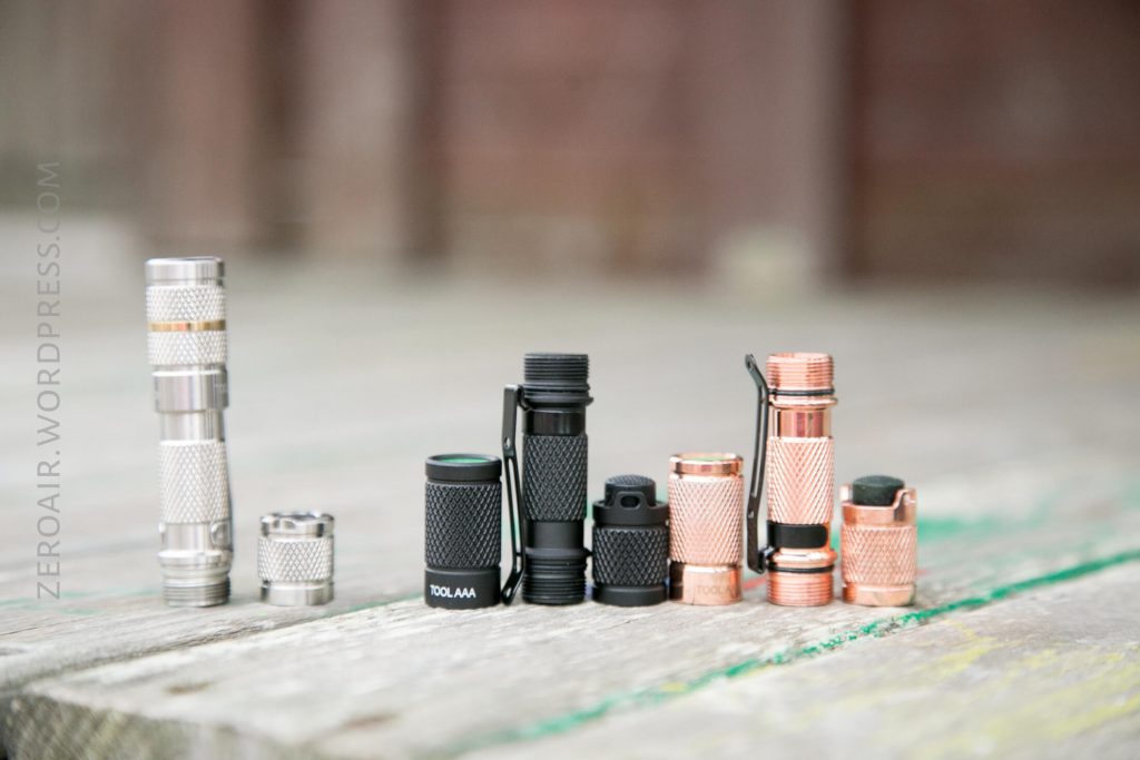 A row of seven small flashlights with different finishes, including silver, black, and copper, are lined up on a weathered wooden surface. A watermark on the left reads ZEROAIR.WORDPRESS.COM.