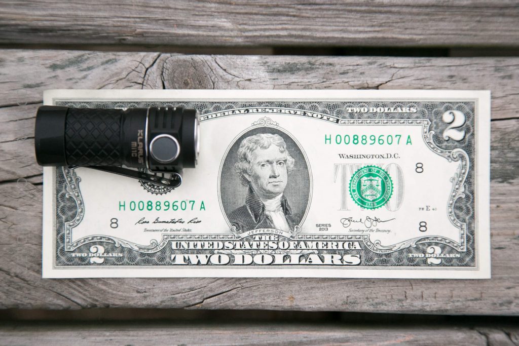 A black flashlight is placed on the left side of a two-dollar bill featuring Thomas Jefferson, which is lying flat on a weathered wooden surface.