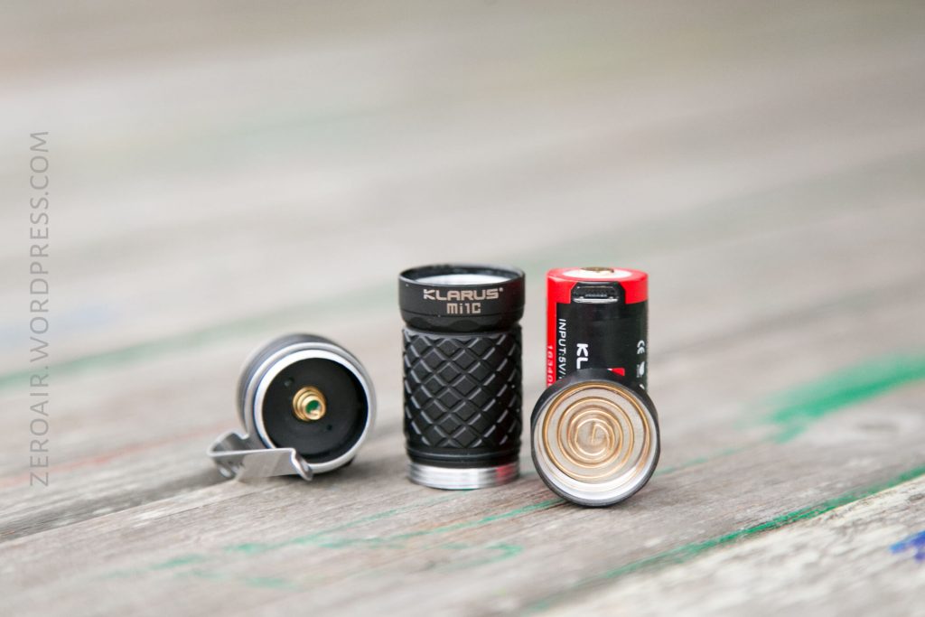 A disassembled Klarus Mi1C flashlight, showing the head, body, and a red Klarus 16340 rechargeable battery, all placed upright on a wooden surface.