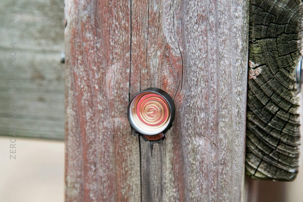 A metal peephole is installed in a weathered wooden door. The wood has a rough texture and visible grain, with some discoloration and cracks around the peephole.