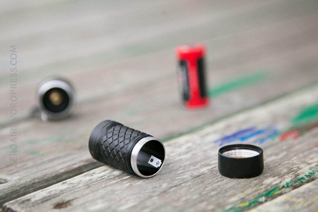 A small black cylindrical device with a textured grip is unscrewed on a wooden surface, showing its metal interior. Three batteries, one red and two black, are out of focus in the background.