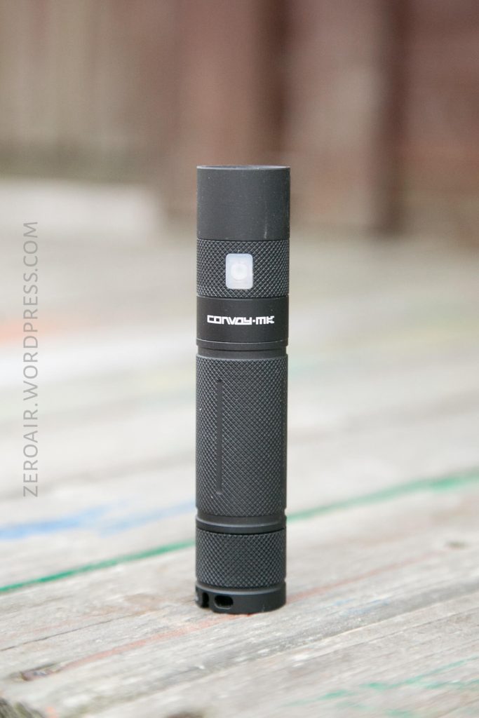 A black Convoy MK flashlight stands upright on a wooden surface with a blurred background. The flashlight has a textured grip and a side button. The text ZEROAIR.WORDPRESS.COM is visible along the left side.