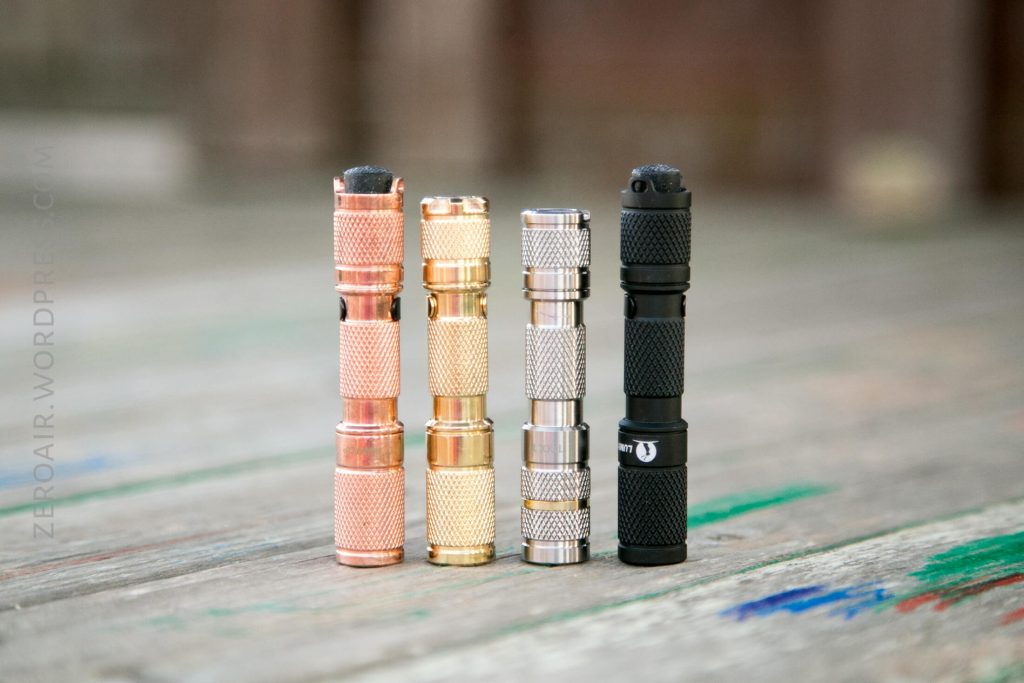 Four compact flashlights are standing upright in a row on a wooden surface. The flashlights are different colors: copper, brass, silver, and black, each with a textured grip pattern.