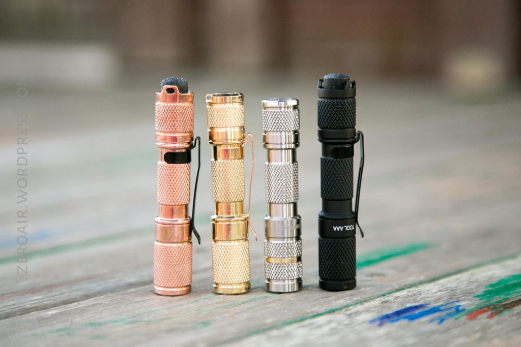 Four small flashlights in copper, gold, silver, and black finishes stand upright on a wooden surface with blurred background. Each flashlight has a textured grip and a button on top.
