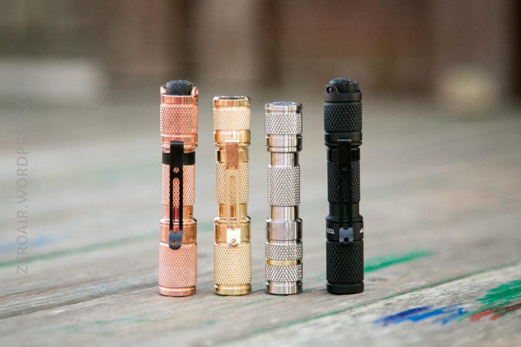 Four small flashlights stand upright on a wooden surface, each with a textured grip. From left to right, the finishes are copper, brass, stainless steel, and black. The background is blurred.