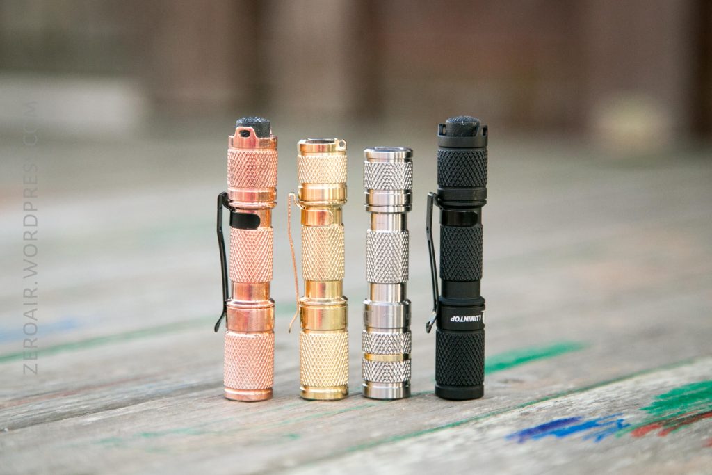 Four small flashlights stand upright in a row on a wooden surface. The flashlights are colored copper, gold, silver, and black, each with a textured grip and pocket clip. The background is blurred.