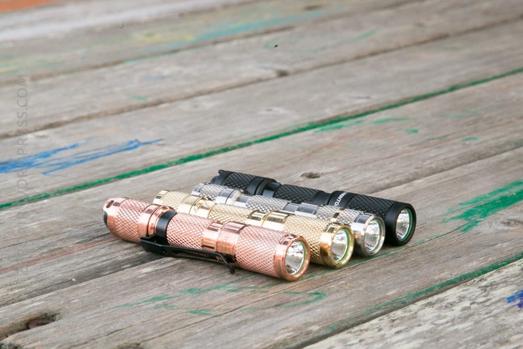 Four metal flashlights in different colors—copper, gold, silver, and black—are placed side by side on a weathered wooden surface. They are aligned in a row with their lenses facing forward.