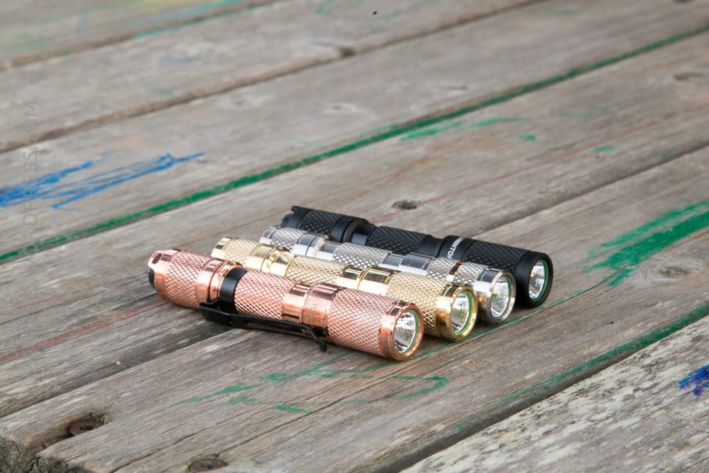 Four compact flashlights in rose gold, silver, gold, and black are lined up on a weathered wooden surface with faint blue and green markings. The flashlights are arranged side by side, facing the same direction.