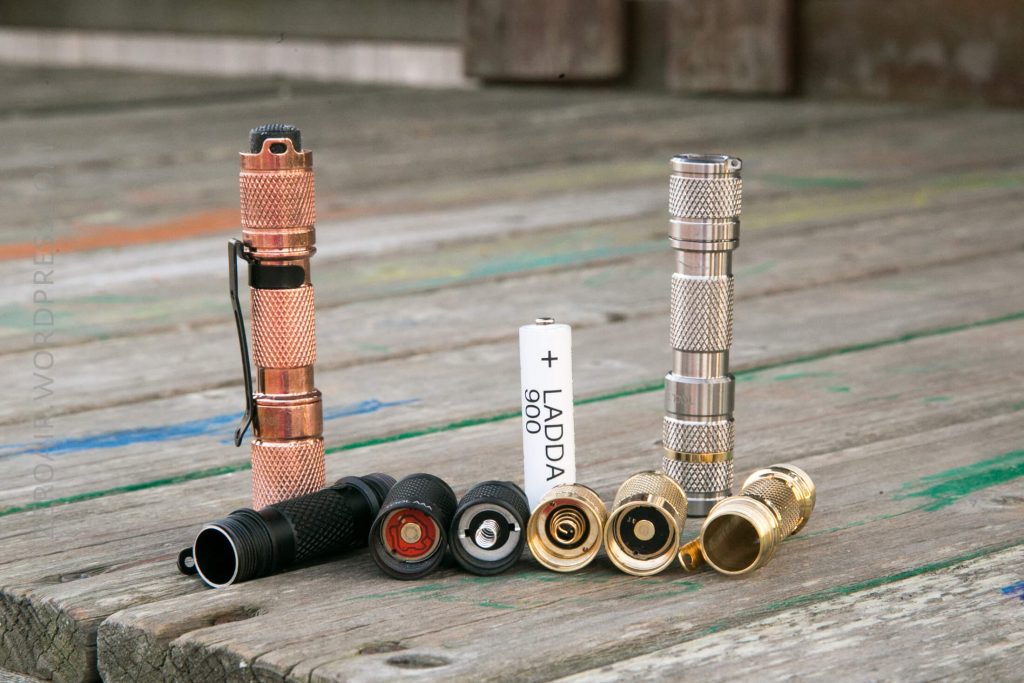Three metal flashlights, one copper-colored, one silver, and one gold, are shown disassembled on a wooden surface with their battery compartments and an AA rechargeable battery visible.