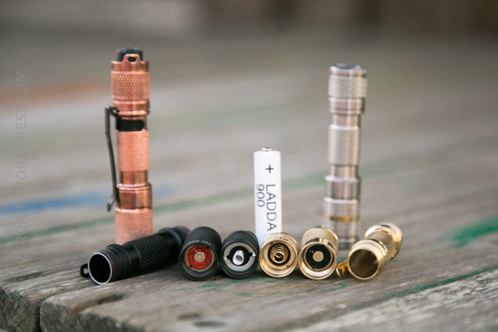 Two flashlights, one copper and one silver, are shown partially disassembled on a wooden surface. The battery compartments and end caps are separated, with one battery marked LADDA 900 displayed in between.