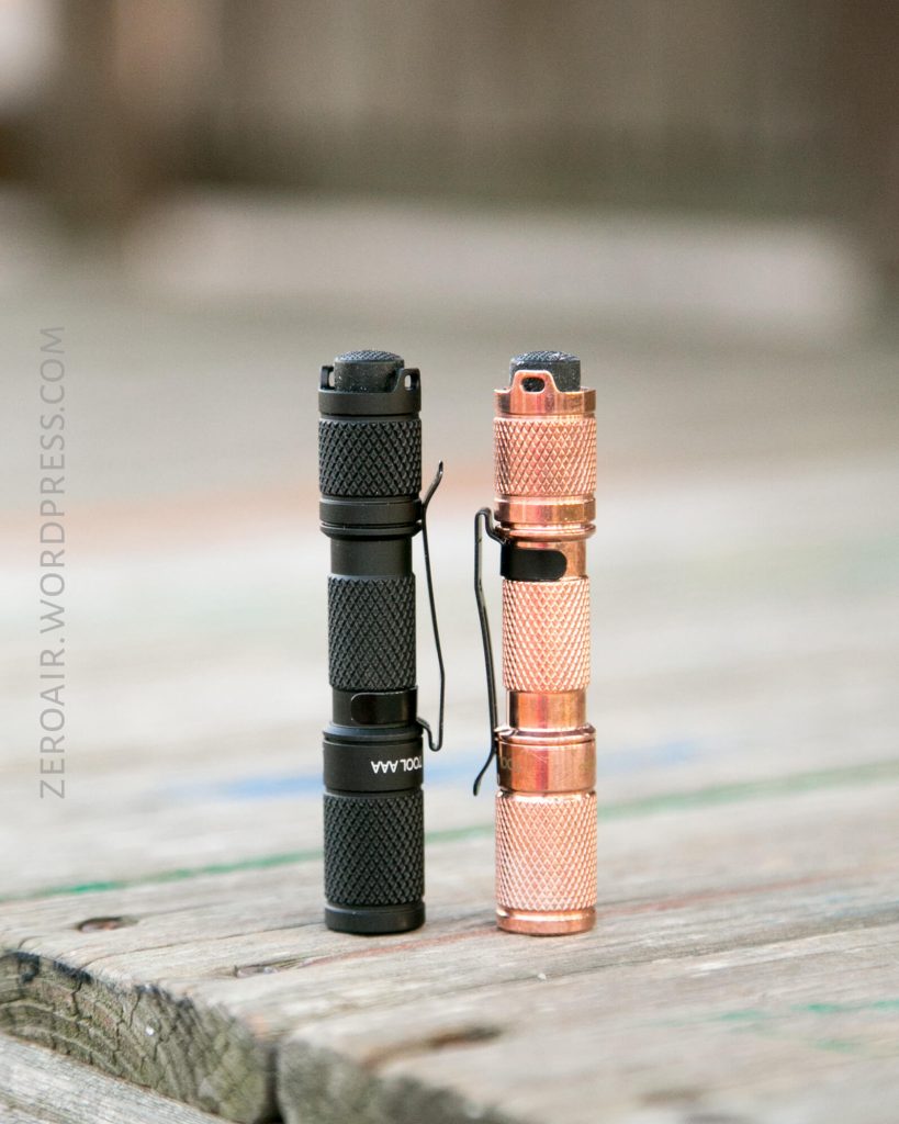 Two small flashlights, one black and one copper-colored, stand upright side by side on a wooden surface. Both have textured grips and metal clips. The background is out of focus.