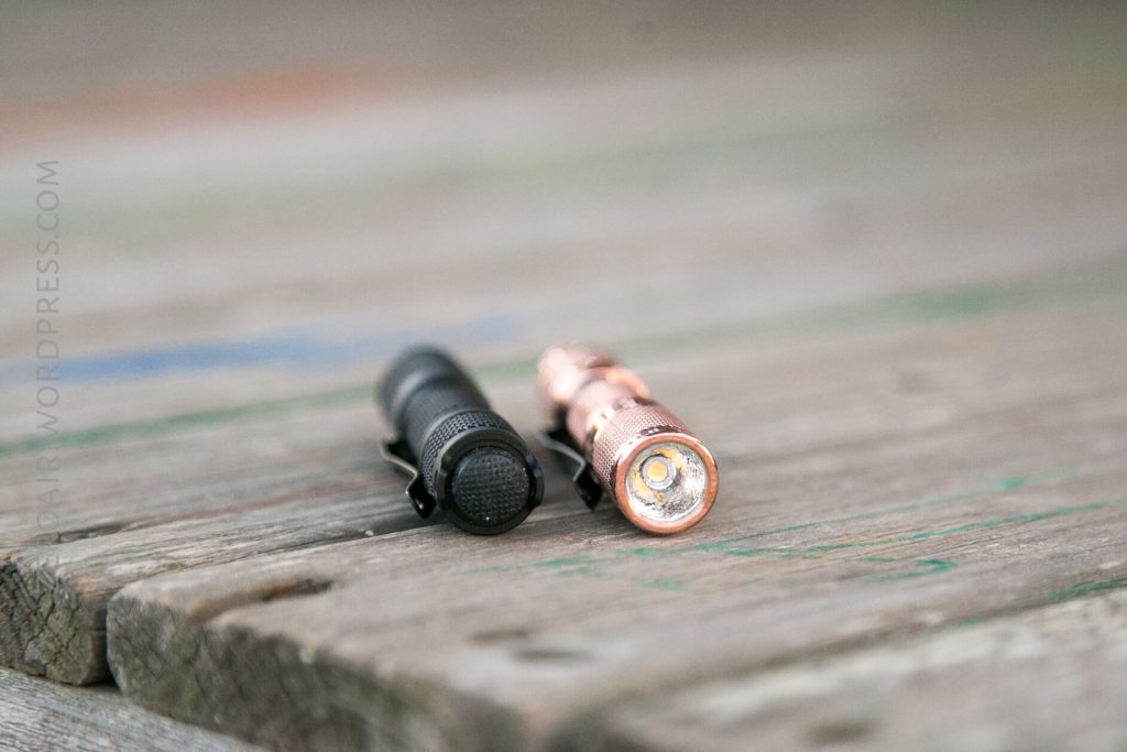 Two small flashlights, one black and one copper-colored, are lying side by side on a weathered wooden surface. The black flashlight is shown from the rear, and the copper one is shown from the front.