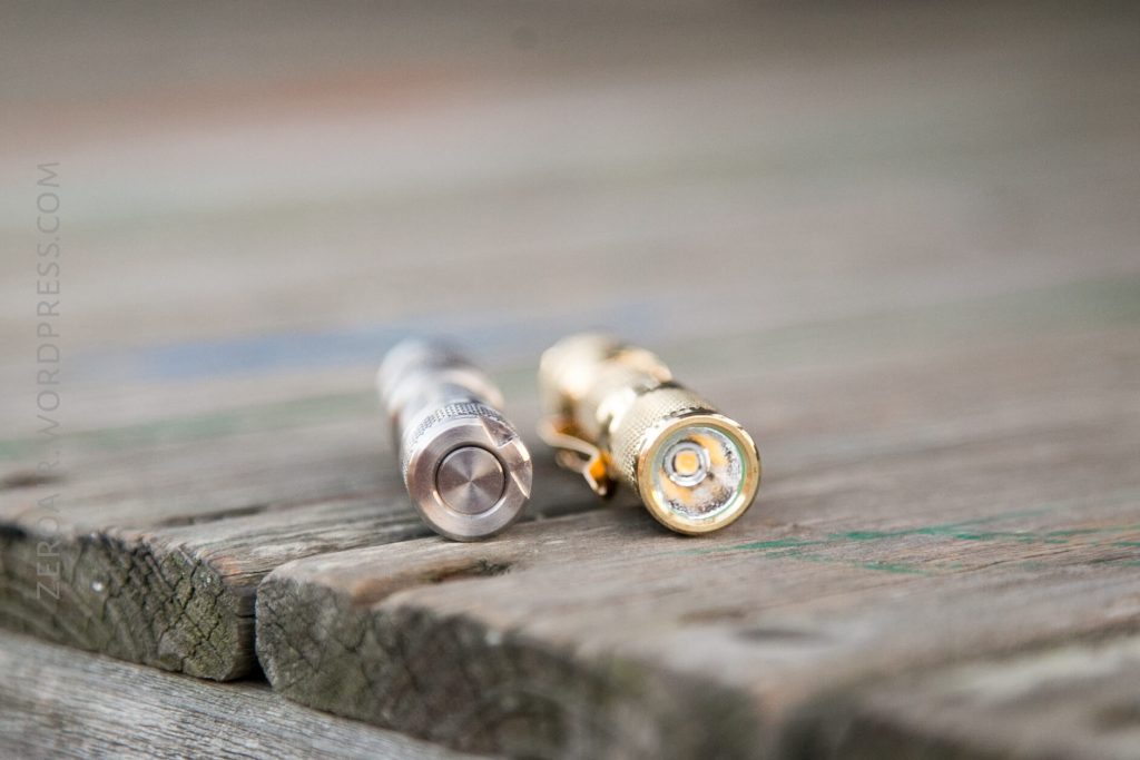 A small stainless steel flashlight and a brass flashlight are lying on their sides on a weathered wooden surface, with the buttons and lenses facing the camera.