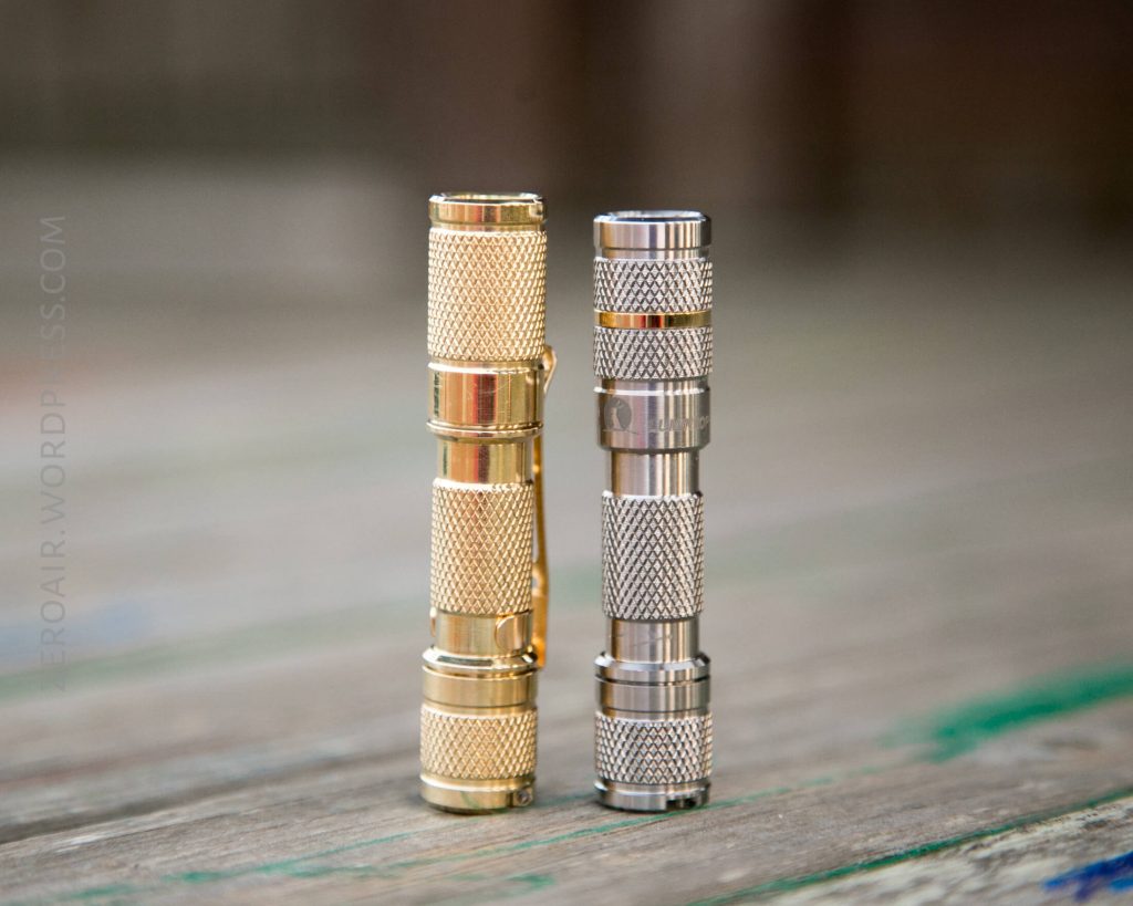 Two small cylindrical flashlights, one gold and one silver, stand upright on a wooden surface. The flashlights have a textured grip and metallic finish.