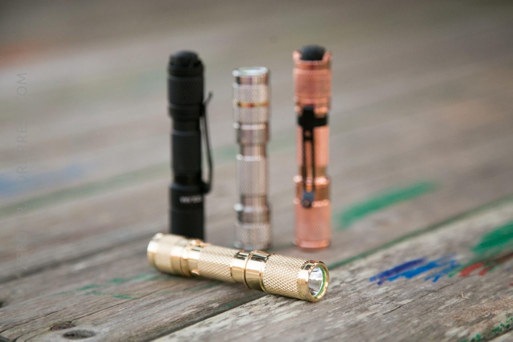 Four small, cylindrical flashlights in black, silver, copper, and gold colors are standing on a weathered wooden surface, with the gold flashlight lying horizontally in the front and the others standing vertically in the background.