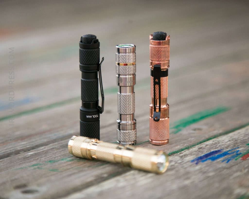 Four compact metal flashlights are standing upright on a wooden surface, each with a different color and finish: black, silver, copper, and gold. One gold flashlight lies horizontally in front of the others.
