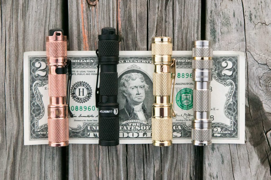 Four metal flashlights in copper, black, gold, and silver are lined up on top of a two-dollar bill on a weathered wooden surface. The flashlights are similar in size and design.