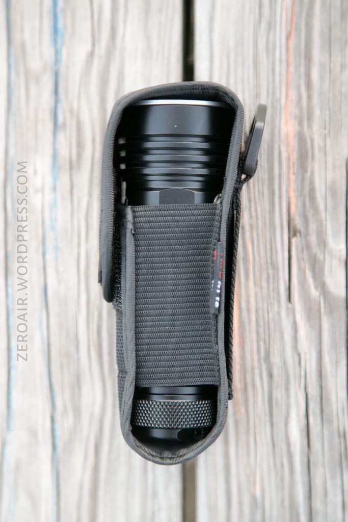 A black flashlight in a partially open dark gray fabric holster is placed vertically on a weathered wooden surface. The zipper of the holster is slightly open, revealing the top of the flashlight.